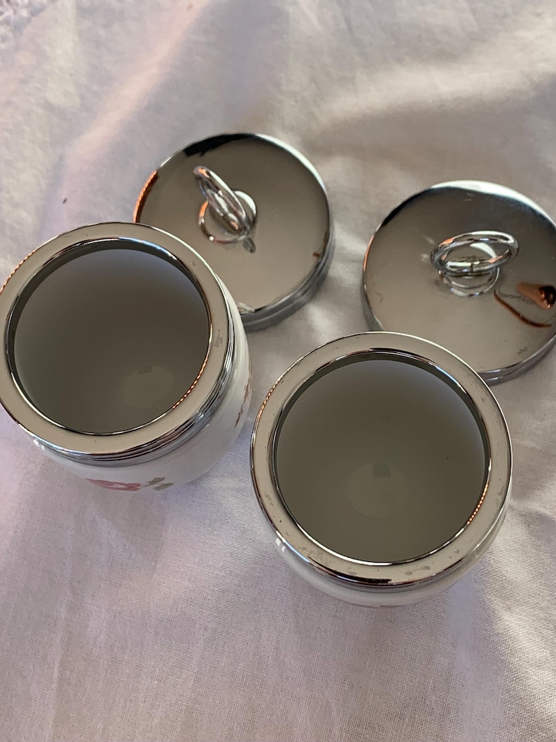 May include: Two white ceramic jars with silver-toned metal rims and matching circular lids. Each lid features a small, upright silver-toned ring handle. The jars have a smooth, white interior and a floral pattern visible on the exterior side.