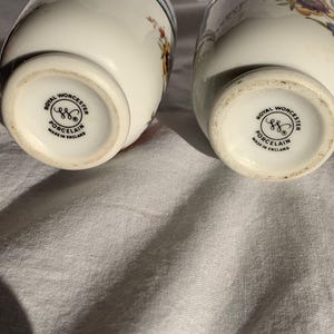 May include: Two white porcelain egg coddlers featuring colorful floral patterns and silver-tone metal lids. The base of each piece is stamped with the Royal Worcester logo, the text Porcelain, and Made in England, indicating their origin.