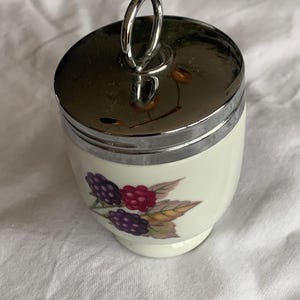 May include: A small, cylindrical ceramic egg coddler with a white base and a polished chrome metal lid featuring a vertical ring handle. The body of the coddler is decorated with a botanical illustration of purple and red blackberries with green leaves.