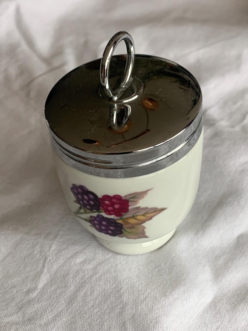 May include: A small, cylindrical ceramic egg coddler with a white base and a polished chrome metal lid featuring a vertical ring handle. The body of the coddler is decorated with a botanical illustration of purple and red blackberries with green leaves.