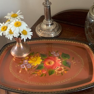 May include: An oval-shaped tray with a reddish-brown finish, featuring a hand-painted floral design in the centre with yellow, red, and purple flowers. The tray has a black border with gold-coloured decorative accents along the outer edge.