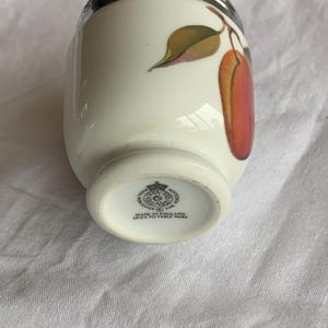 May include: A white porcelain egg coddler with a silver-colored metal lid. The body features a painted design of a red fruit with green leaves. The base is stamped with the Royal Worcester logo and the text Made in England Oven to Table Ware.