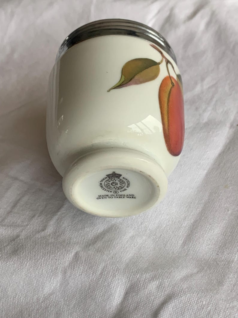 May include: A white porcelain egg coddler with a silver-colored metal lid. The body features a painted design of a red fruit with green leaves. The base is stamped with the Royal Worcester logo and the text Made in England Oven to Table Ware.