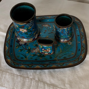 May include: A desk set featuring a rectangular tray with rounded corners and three cylindrical containers of varying heights. The set is crafted from blue metal with intricate floral patterns in red, green, white, and yellow, finished with dark metal rims.