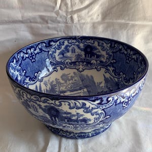May include: A large ceramic bowl featuring a blue and white transferware pattern. The exterior and interior display detailed landscape scenes with architectural ruins, surrounded by ornate scrollwork and decorative floral borders.