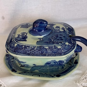 May include: A square ceramic gravy boat with a matching lid and underplate, featuring a blue and white willow pattern. The piece includes a dark blue handle and a knob on the lid, decorated with detailed landscape scenes of bridges and trees.