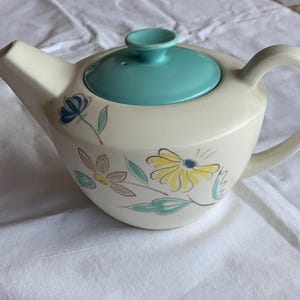 Può includere: Una teiera in ceramica color crema con un coperchio turchese brillante e un manico. Il corpo della teiera è decorato con motivi floreali dipinti a mano nei toni del giallo, blu e turchese, che mostrano delicati petali e foglie.