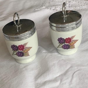 May include: Two white ceramic egg coddlers with shiny silver metal lids and ring handles. Each coddler features a colorful botanical illustration of blackberries and raspberries with green leaves on the front of the rounded body.