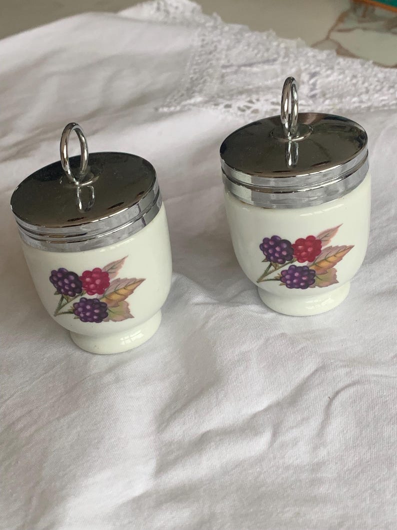 May include: Two white ceramic egg coddlers with shiny silver metal lids and ring handles. Each coddler features a colorful botanical illustration of blackberries and raspberries with green leaves on the front of the rounded body.