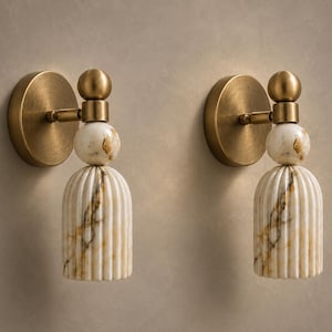May include: A pair of wall sconces featuring a circular brushed brass backplate, a small brass sphere, and a fluted, bell-shaped shade made of white marble with brown veining. Each light fixture includes a spherical marble accent piece.