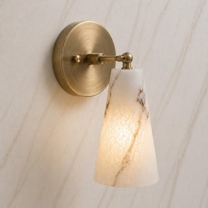 Puede incluir: Aplique de pared con base circular de latón y pantalla cónica. La pantalla es blanca con vetas marrones, y la lámpara está iluminada desde dentro. El aplique está montado en una pared clara.