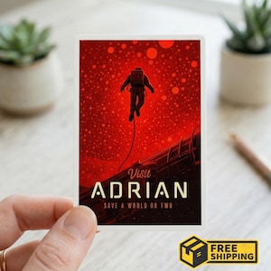 May include: A rectangular sticker featuring an astronaut floating in a red space scene. The text "Visit Adrian" and "Save a World or Two" are displayed in a stylized font. The sticker is held by a hand.