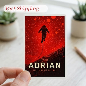 May include: A rectangular sticker featuring an astronaut in a spacesuit against a red background with a dotted pattern. The sticker reads "Visit Adrian" and "Save a World or Two" in a stylized font.