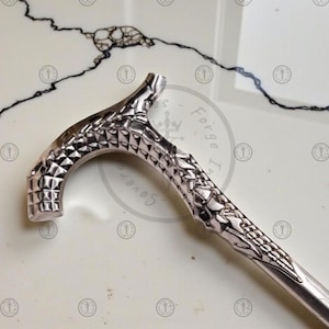 May include: A silver-colored walking cane with a textured handle and shaft. The handle has a geometric pattern, and the shaft tapers to a point. The cane is designed for support and style.
