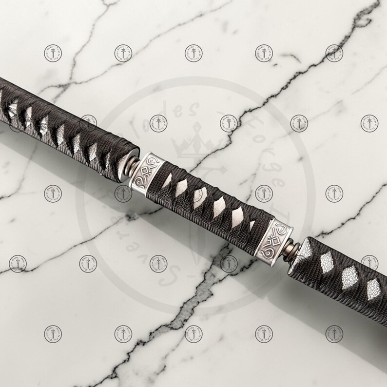 May include: Close-up of a katana handle and blade. The handle is wrapped in black fabric with white diamond-shaped accents. Silver-colored metal accents and fittings are visible. The blade is partially visible, with a black and silver design.