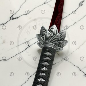 May include: A katana sword with a red blade and a silver lotus flower-shaped handguard. The handle is wrapped in black fabric. The sword is displayed on a white marble surface.