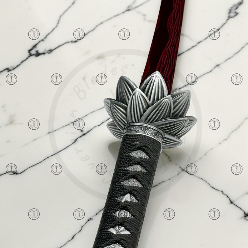 May include: A katana sword with a red blade and a silver lotus flower-shaped handguard. The handle is wrapped in black fabric. The sword is displayed on a white marble surface.