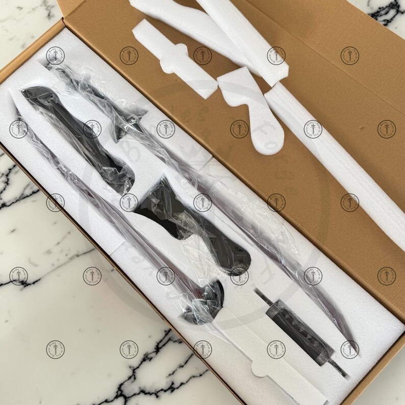May include: A set of black kitchen knives and utensils, including a carving knife, a bread knife, and a fork, packaged in a cardboard box with protective foam inserts. The knives have black handles and silver blades.