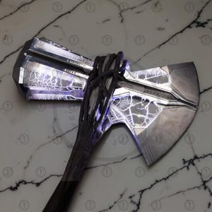 May include: A silver and black battle axe with a detailed design. The axe head has a lightning-like pattern with a dark handle wrapped in a textured material. The axe is a prop or collectible.