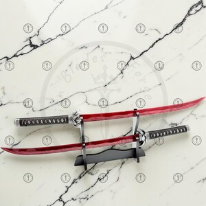 May include: Two katana swords with red blades and black handles are displayed on a black stand against a marble background. The swords have silver accents and intricate handle designs. The blades are curved and appear sharp.