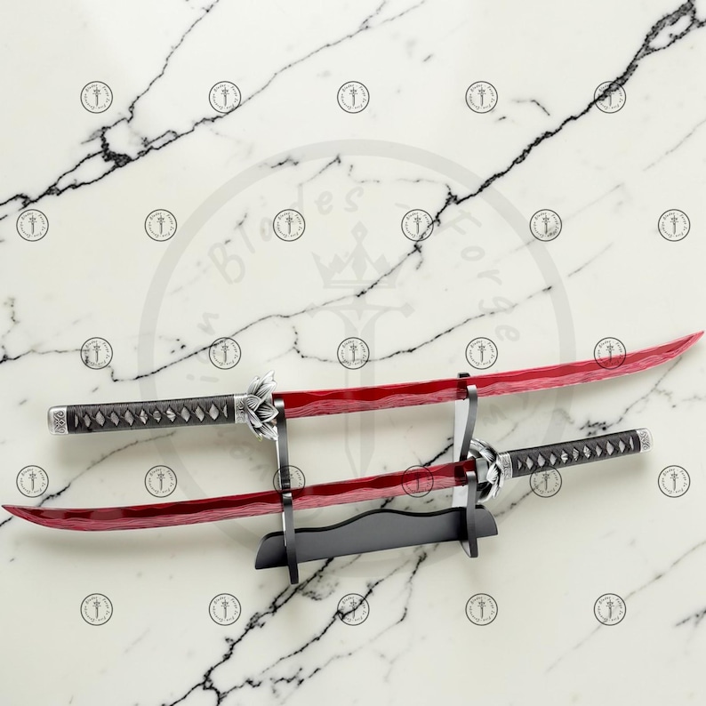 May include: Two katana swords with red blades and black handles are displayed on a black stand against a marble background. The swords have silver accents and intricate handle designs. The blades are curved and appear sharp.