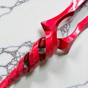 May include: A vibrant red spear with a spiraling shaft and sharp, angular tips. The glossy finish reflects light, highlighting the weapon's intricate design. The spear is set against a white and gray patterned background.
