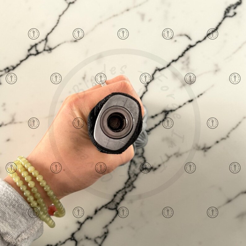 May include: A black and silver bottle stopper with a textured grip. The stopper has a circular design and is held in a hand. The background is a white marble pattern.