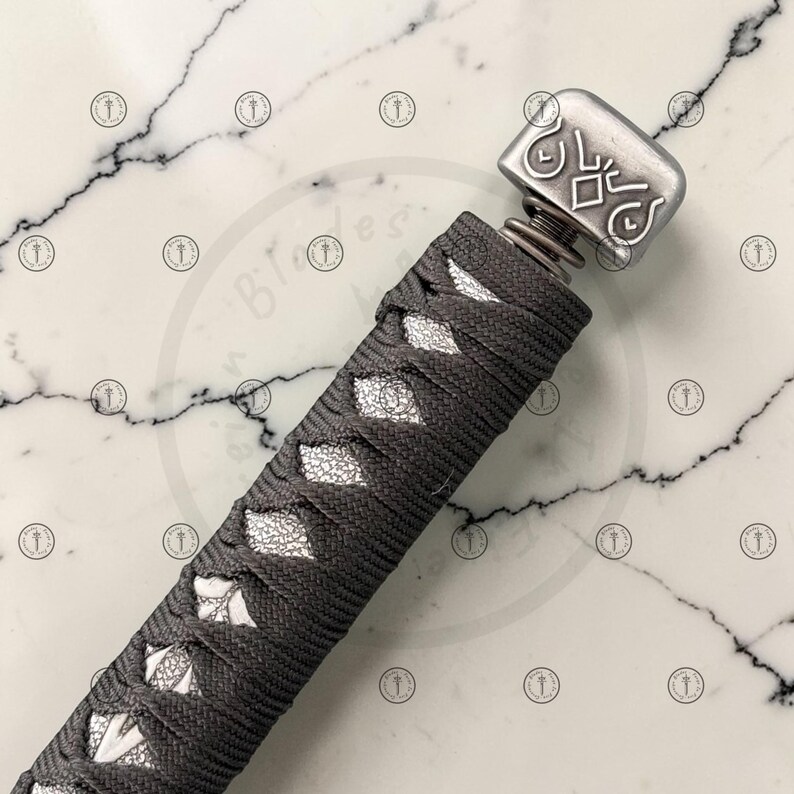 May include: Close-up of a katana handle. The handle is wrapped in dark gray cord with diamond-shaped white accents. The metal pommel is rectangular with engraved designs. A spring is visible between the handle and the pommel.