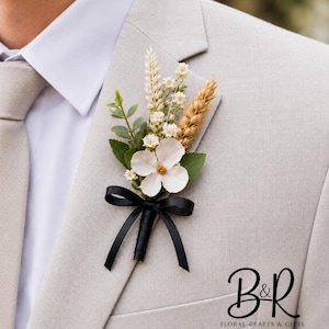 May include: A boutonniere featuring a white flower, green leaves, small white blossoms, and a stalk of wheat, all tied together with a black ribbon. The floral arrangement is pinned to a light beige suit jacket. Text B&R Floral Crafts & Gifts.