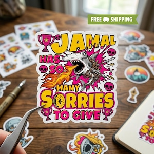 May include: A sticker with the text "JAMAL HAS SO MANY SORRIES TO GIVE" in yellow and pink. The design features a shark with mechanical elements breathing fire, surrounded by skulls and trophies. Other stickers are visible on a wooden surface.