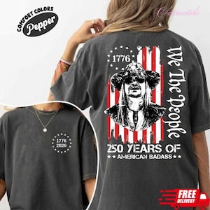 May include: Dark gray t-shirt with a graphic of a man wearing a hat and sunglasses, set against a red, white, and blue flag design. The text reads "1776" and "We The People", with "250 YEARS OF AMERICAN BADASS" below. A smaller design with "1776 2026" is on the front.
