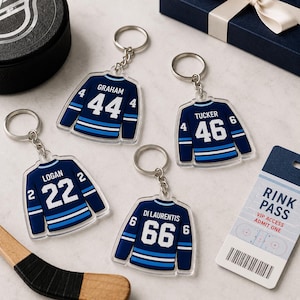 May include: Four acrylic keychains shaped like dark blue hockey jerseys with white stripes and numbers. The jerseys feature the names GRAHAM 44, TUCKER 46, LOGAN 22, and DI LAURENTIS 66. Each keychain includes a silver metal ring and chain attachment.