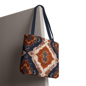May include: Tote bag with a colorful, geometric pattern in shades of orange, navy blue, and cream. The bag has navy blue handles and is made of a durable fabric. The design resembles a traditional rug.