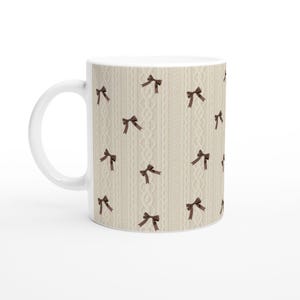 Op de afbeelding: Witte keramische koffiemok met een herhalend patroon van bruine strikken op een crèmekleurige achtergrond. De achtergrond heeft een kabelbreiselontwerp. De mok heeft een wit handvat en een glanzende afwerking.