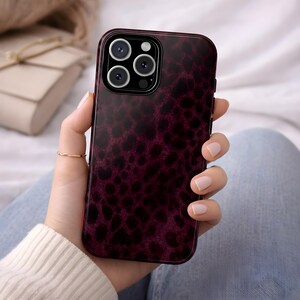 May include: A burgundy phone case with a leopard print design. The case has a glossy finish and features a camera module with multiple lenses. The phone case is held in a person's hand.