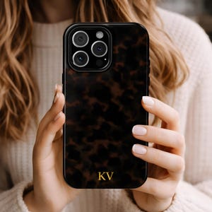 May include: A black phone case with a tortoiseshell pattern is held by a person. The case has a gold monogram of the letters "KV" at the bottom. The phone case has a black border and a camera module with three lenses.