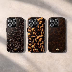 May include: Three mobile phone cases with a tortoiseshell design in brown and gold tones. Each case features a gold initial: LK, KA, and AD. The cases have a shiny finish and a camera aperture.