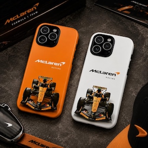 May include: Two smartphone cases featuring the McLaren Racing logo and a graphic of a Formula 1 race car. One case is bright orange and the other is white. Both cases have a black camera cutout area and display the McLaren Racing text.