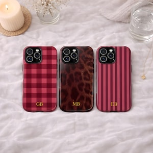 May include: Three phone cases with different designs. The first is a red and burgundy plaid pattern with the letters "GB". The second has a brown tortoiseshell pattern with the letters "MB". The third has burgundy vertical stripes with the letters "EB".