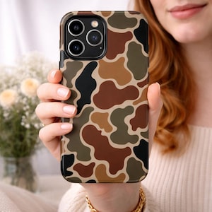 May include: A phone case with a camouflage pattern in shades of brown, black, and tan. The case has a glossy finish and is held by a person. The phone case has a camera module with three lenses.