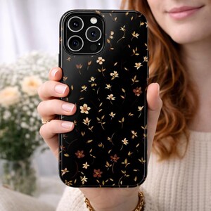 May include: Black phone case with a floral pattern. The case features a repeating design of small, cream and brown flowers and leaves. The phone case is held in a person's hand.
