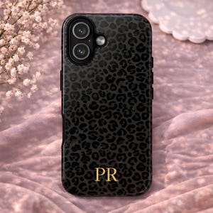 以下が含まれることがあります： 黒いスマートフォンケース。ヒョウ柄模様と金色のイニシャル「PR」が特徴です。ケースにはカメラ用の切り抜きがあり、白い花のあるピンク色の背景に置かれています。
