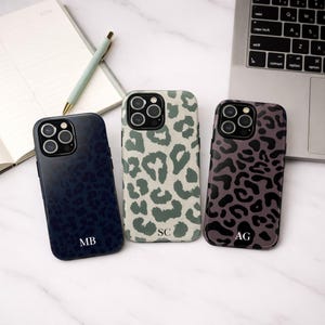 以下が含まれることがあります： ヒョウ柄のデザインの携帯電話ケースが3つ。1つはイニシャル「MB」入りのネイビーブルー、もう1つは「SC」入りのグリーンと白、3つ目は「AG」入りの紫と黒。ケースは白い表面に置かれています。