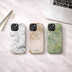 Puede incluir: Tres fundas para teléfonos inteligentes con diseños únicos. La primera tiene un patrón de mármol blanco con vetas negras y las iniciales "PA". La segunda tiene un diseño de remolino beige y bronceado con las iniciales "DM". La tercera tiene un diseño de remolino verde y blanco con las iniciales "KT".