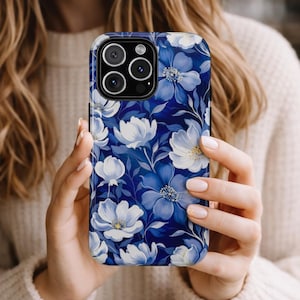 May include: A smartphone case featuring a floral pattern in shades of blue and white. The case has a dark blue background with white and light blue flowers and green leaves. The case is held in a person's hands.