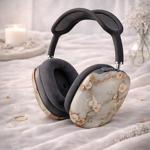Op de afbeelding: Draadloze over-ear hoofdtelefoon met een bloemenontwerp. De oorschelpen hebben een wit en goud marmerpatroon met delicate perzikroze bloemen en takken. De hoofdband en oorschelpen zijn donkergrijs.