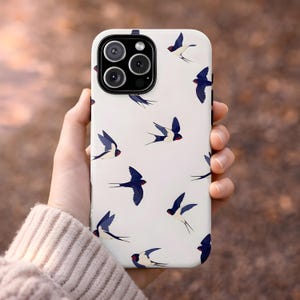 Könnte beinhalten: Weißes Handycase mit einem Muster aus blauen und weißen Schwalben mit roten Akzenten. Das Case hat eine glänzende Oberfläche und einen Kameraausschnitt. Das Handycase wird in einer Hand gehalten.