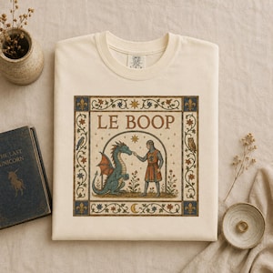 Könnte beinhalten: Ein cremefarbenes T-Shirt mit einem rechteckigen Grafikdruck, der eine mittelalterliche Illustration eines Ritters zeigt, der die Nase eines grünen Drachen berührt. Der Text LE BOOP steht über den Figuren, umrahmt von floralen Mustern.