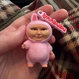 May include: A pink keychain featuring a figure in a bunny suit with a human face. The keychain has the text "lynnbubu" in red. The figure has small arms and feet, and the keychain is attached to a silver ring.