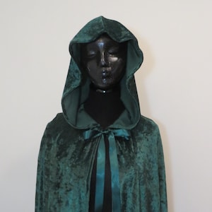 May include: A dark green velvet cloak with a hood and satin ribbon ties. The cloak is displayed on a black mannequin, against a plain white background. The cloak's texture is visible, and the hood is up.